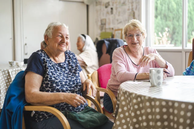 Two elderly ladies sit together around a table, smiling