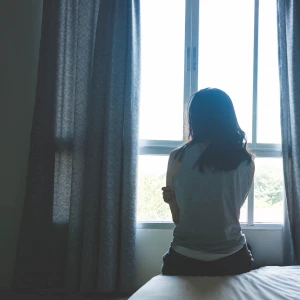An image of a woman sitting on a bed, back to the camera, facing a window.