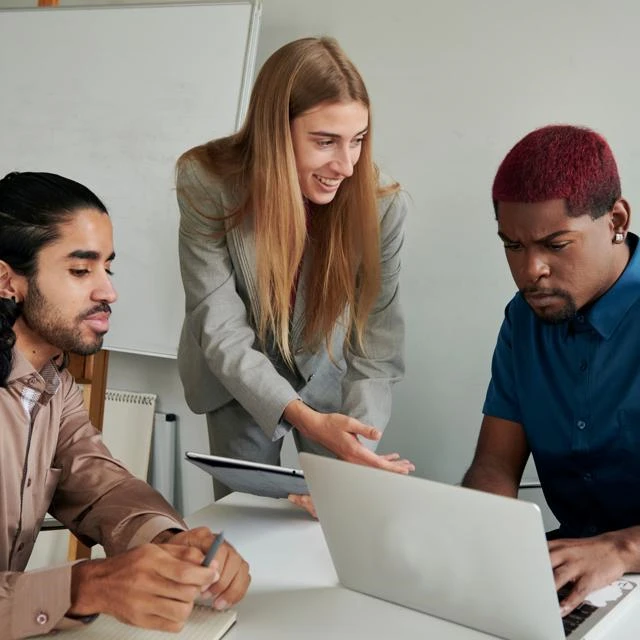 A manager is smiling and showing something on a clipboard to staff whilst they are looking at a screen