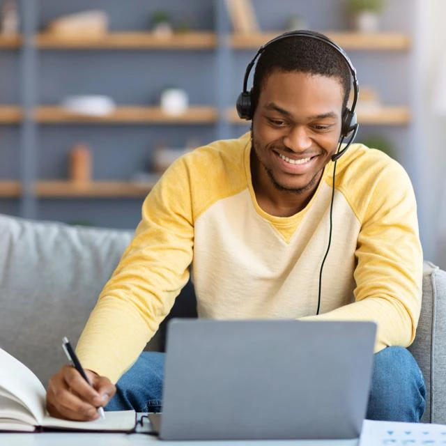 Male learner with headphones and laptop taking notes and smiling