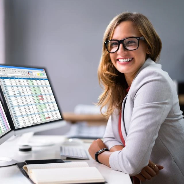a professional woman sitting at a desk with two monitors displaying an EXCEL spreadsheet